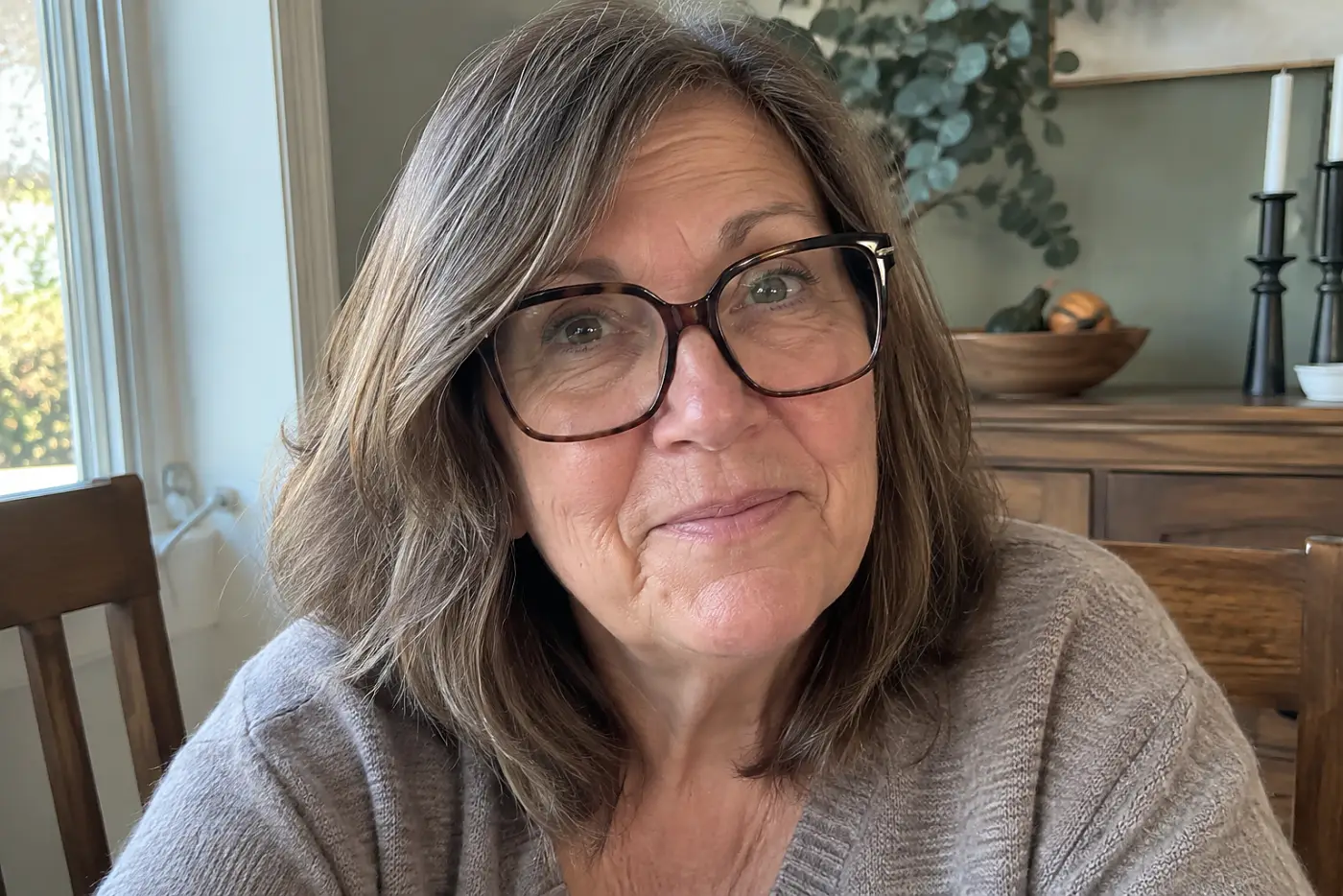A woman in her sixties wears oversized square glasses while sitting at her dining table.