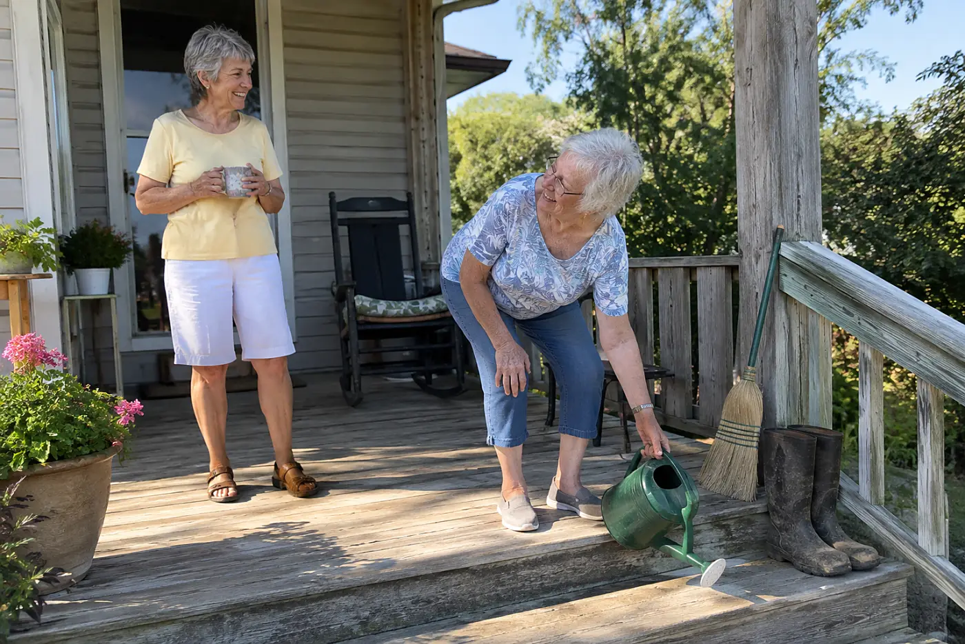 A woman wearing lined bifocals reaches for a watering can while talking with a friend on a porch.