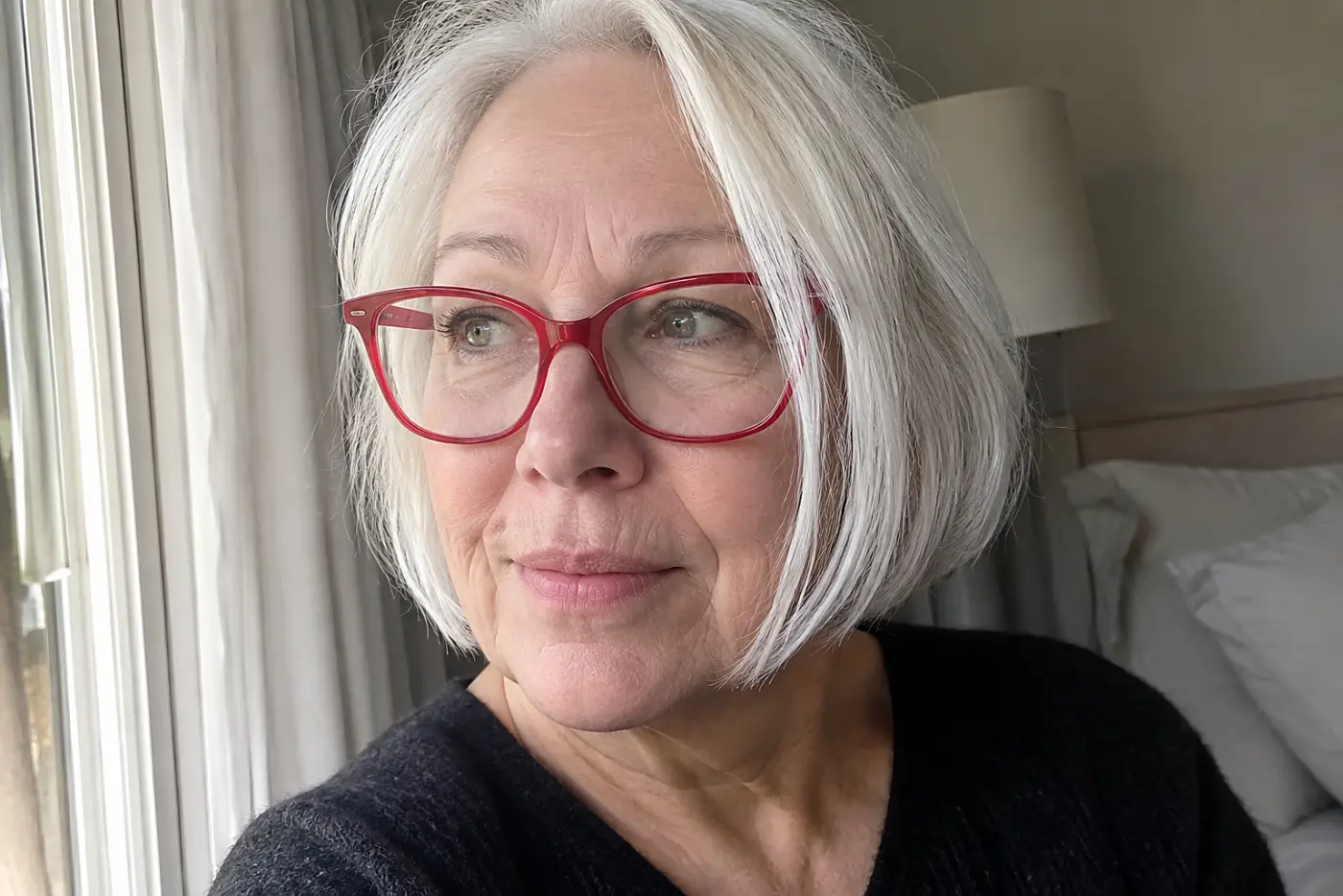 A woman in her sixties with white hair wears bright red glasses beside a window.