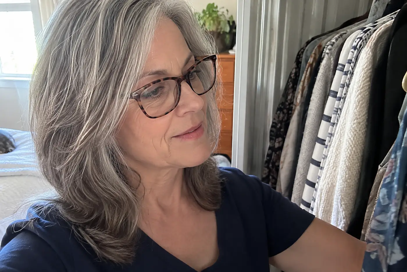 A woman over 50 wearing patterned glasses looks through clothes in her closet.