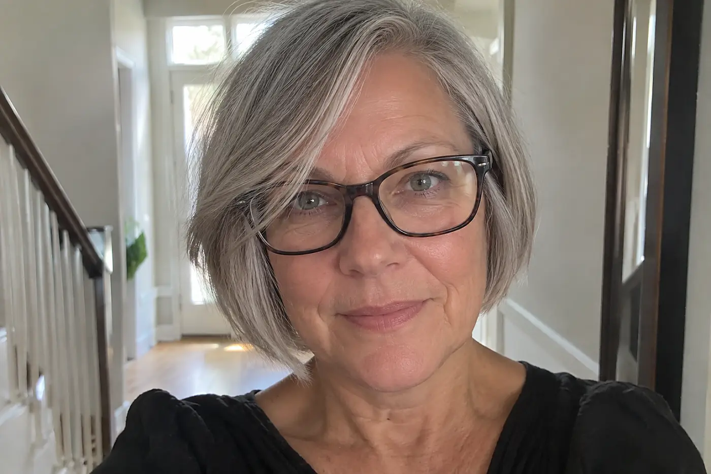 A woman over 50 with short grey hair wears tortoiseshell glasses in a hallway.