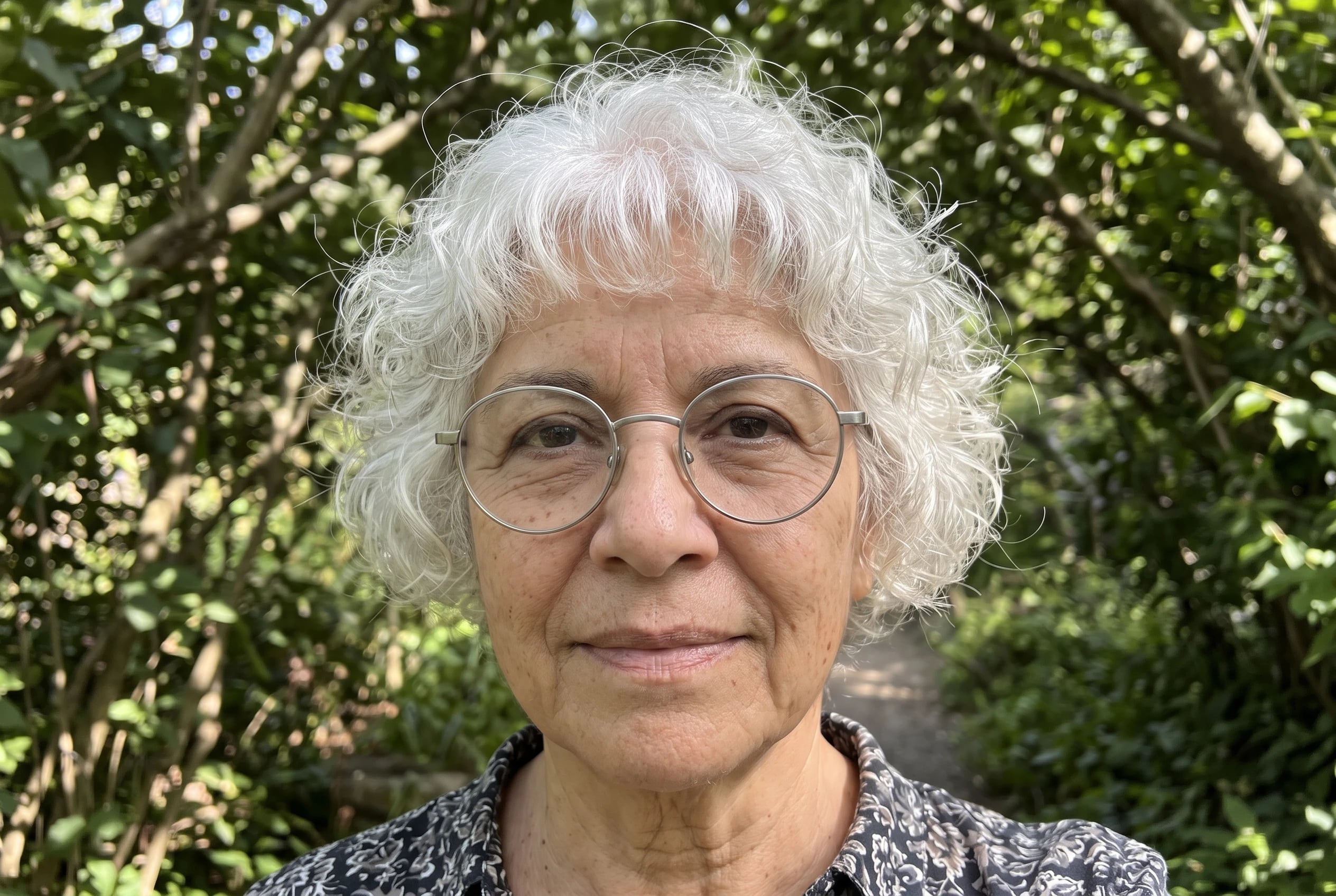 A woman in her sixties with curly white hair wears brushed silver round metal frames outdoors.