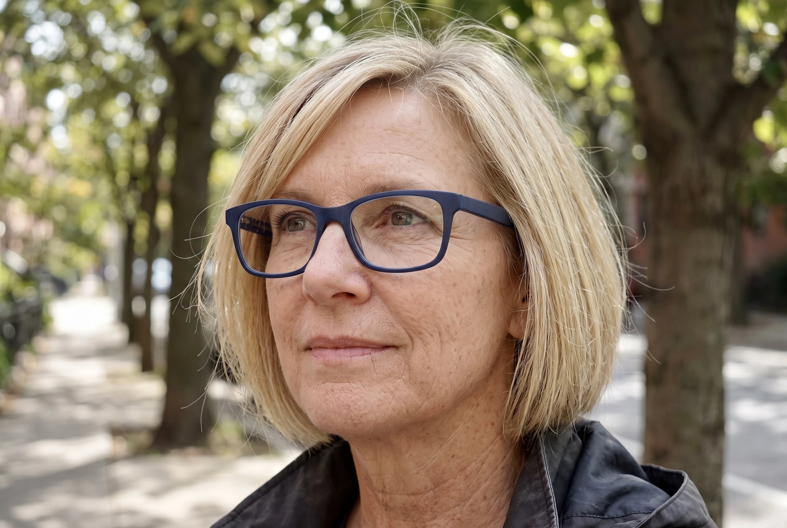 A woman in her fifties wears matte navy rectangle glasses on a tree-lined street.