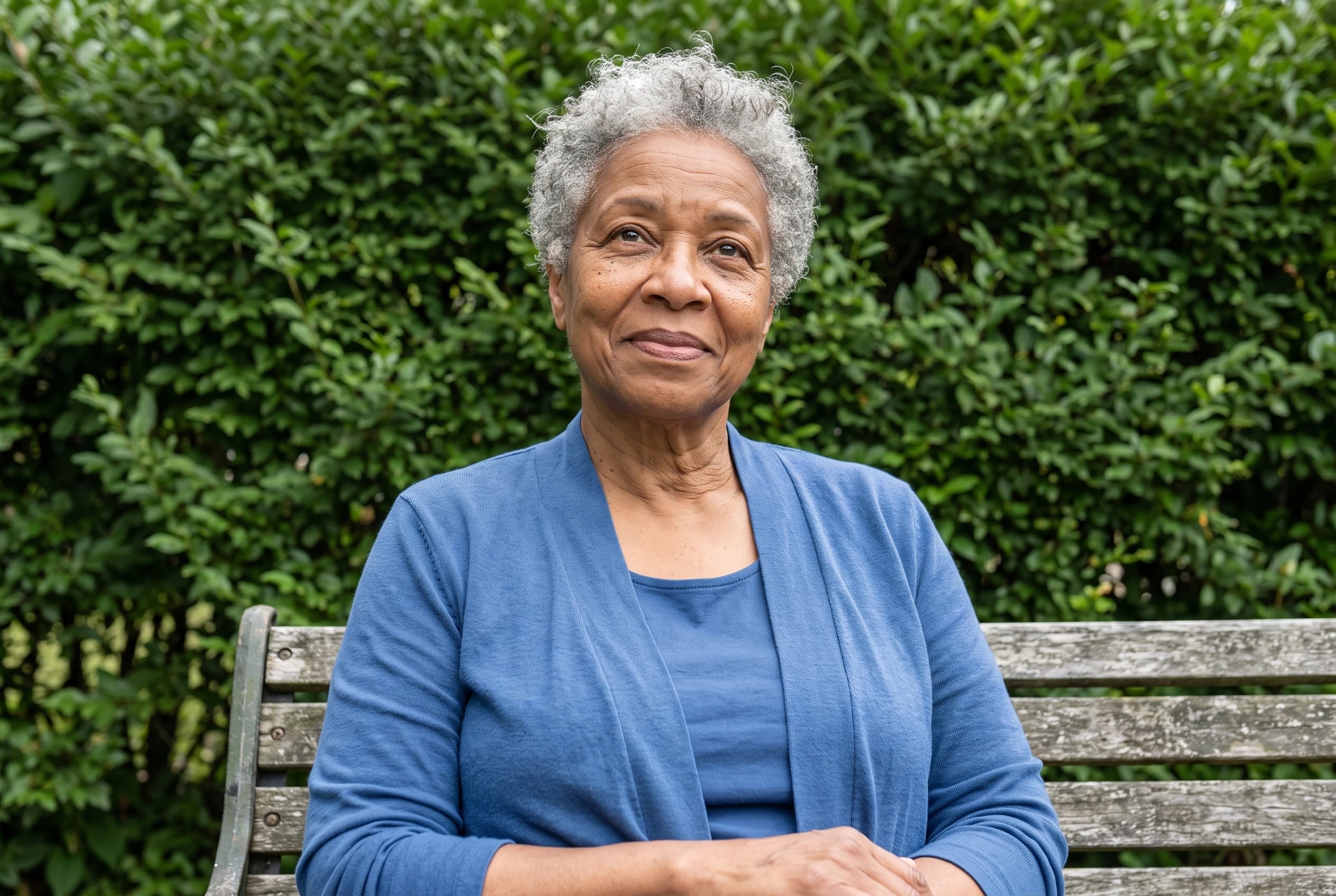 A blue cardigan and matching shell fill the center of the frame on a woman in her sixties sitting on a garden bench.
