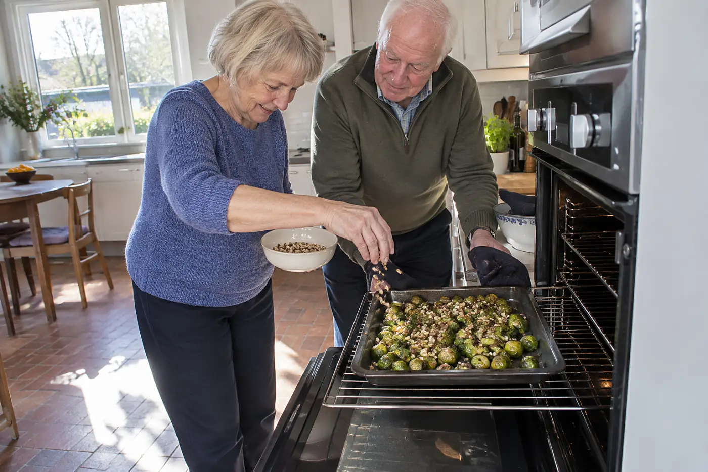 Woman scatters chopped hazelnuts over Brussels sprouts beside an open oven.
