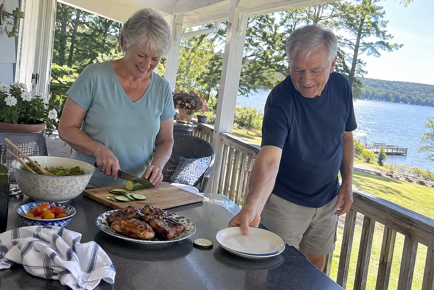 A woman slices cucumber beside glazed chicken on a porch counter.