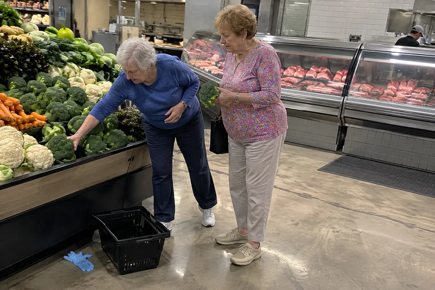 A woman selects broccoli beside the butcher counter at a grocery store.