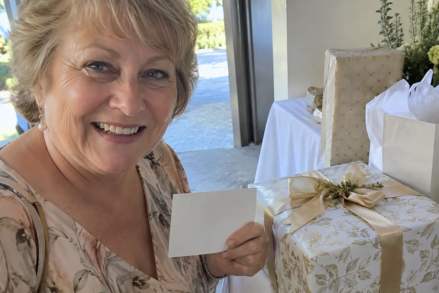 A wrapped box sits near a gift table at a reception entrance as a woman in her sixties holds a small plain card.