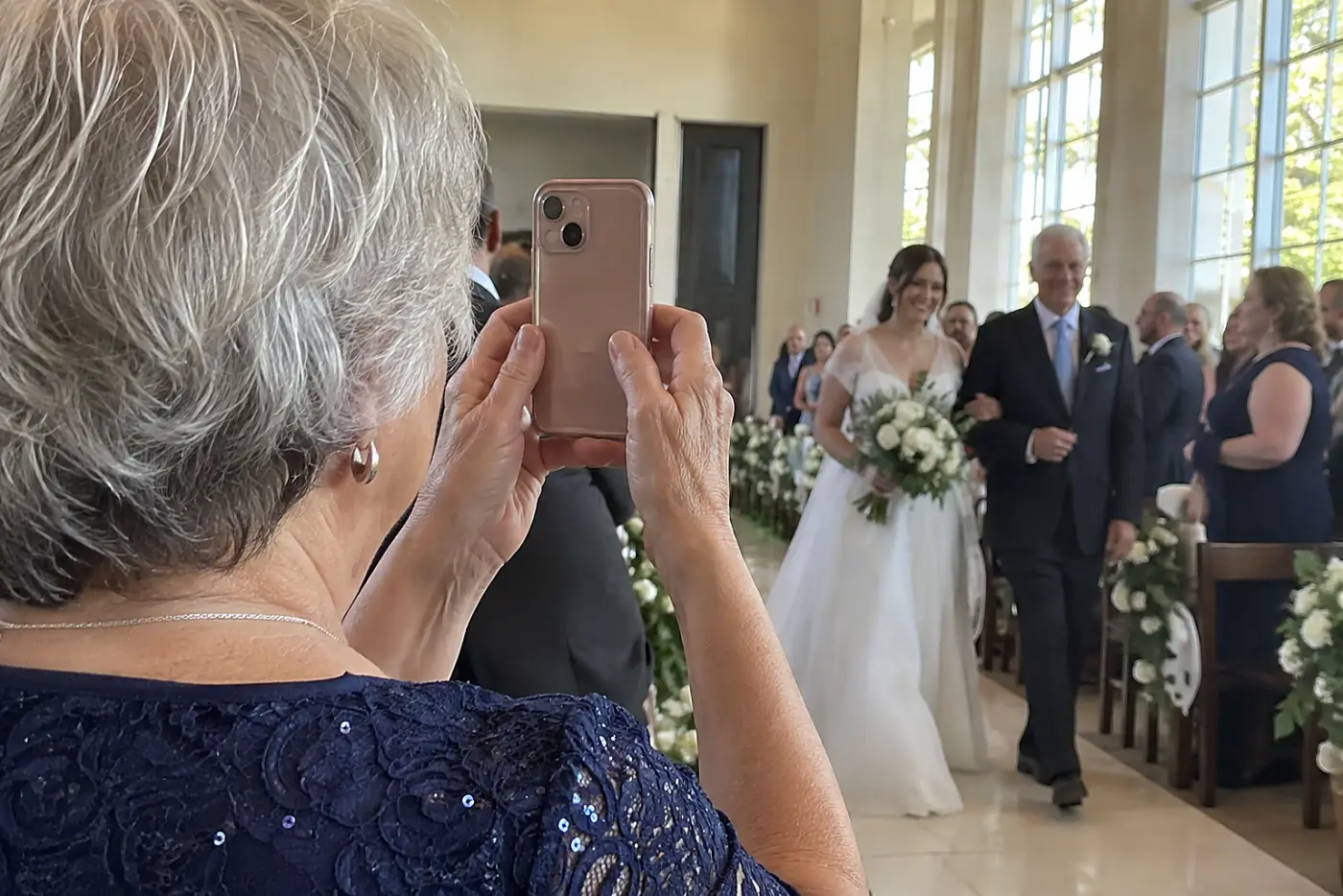 A woman with grey hair holds up a phone while a bride walks down the aisle.