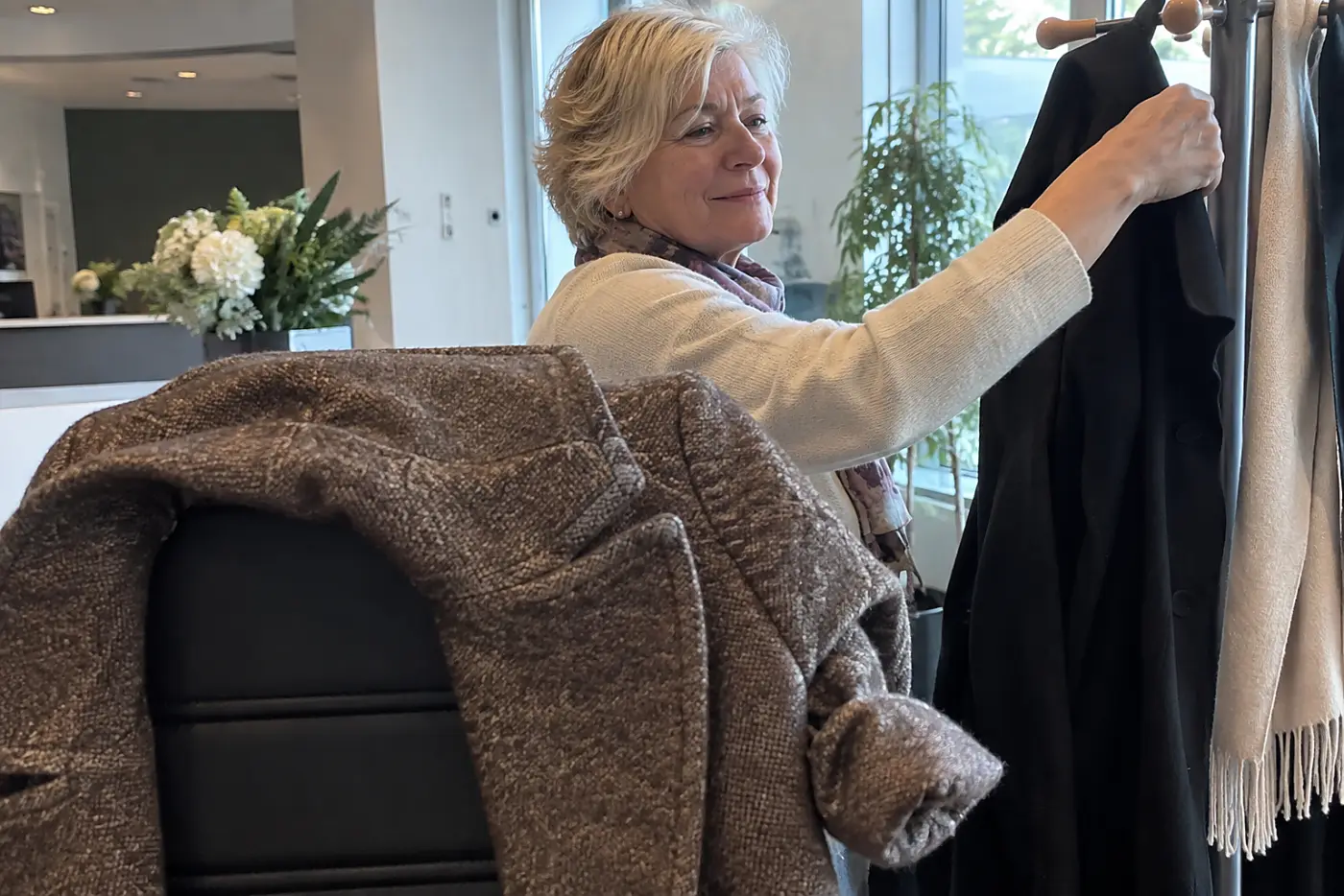 A heavy coat hangs over a reception chair while a woman in her sixties stands beside a nearby coat rack.