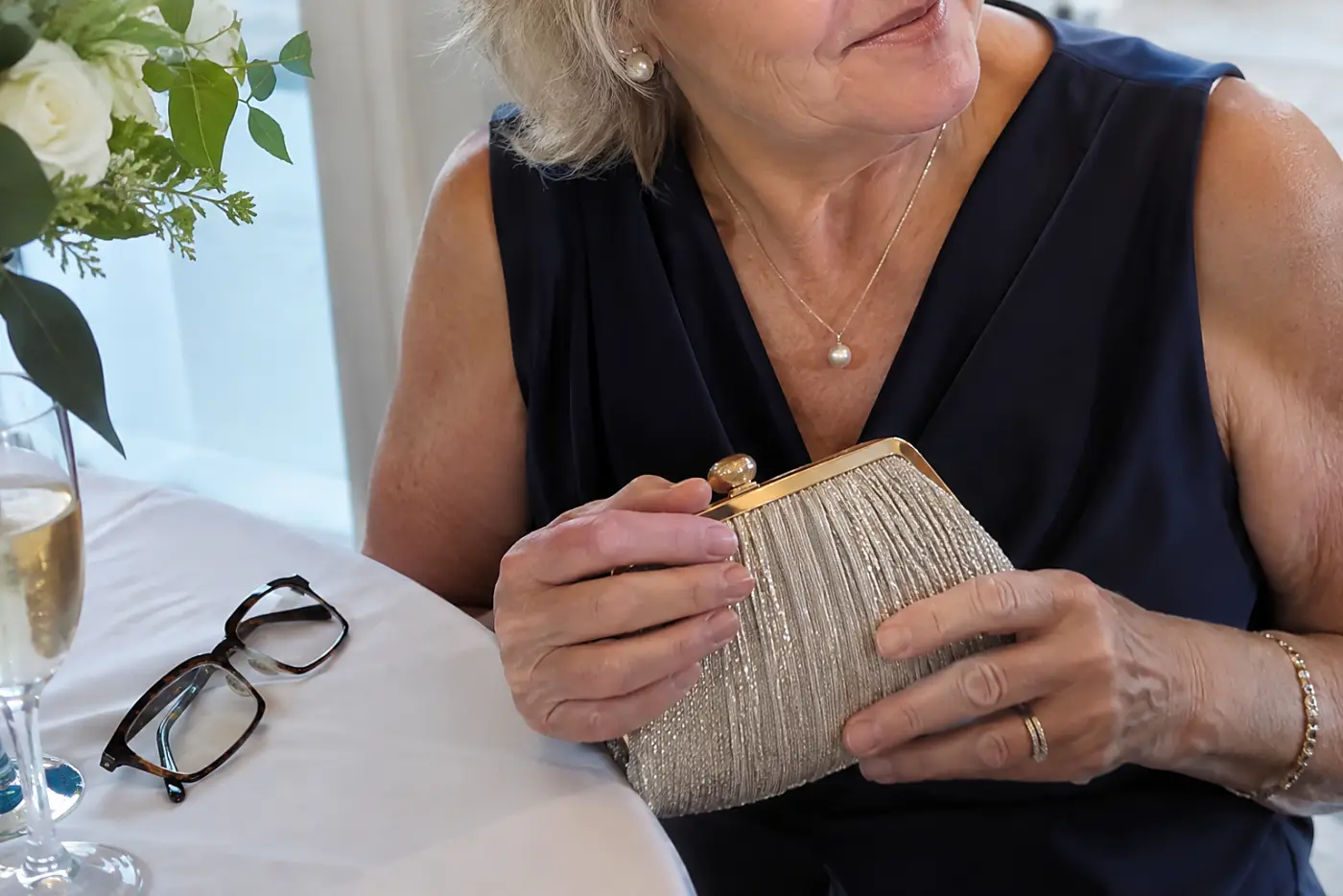 A closed clutch sits in the hands of a woman in her sixties at a reception table, with her glasses beside it.