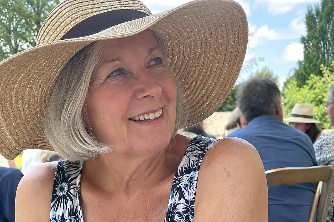 A smiling woman in her sixties wears a wide-brimmed straw hat at an outdoor wedding.