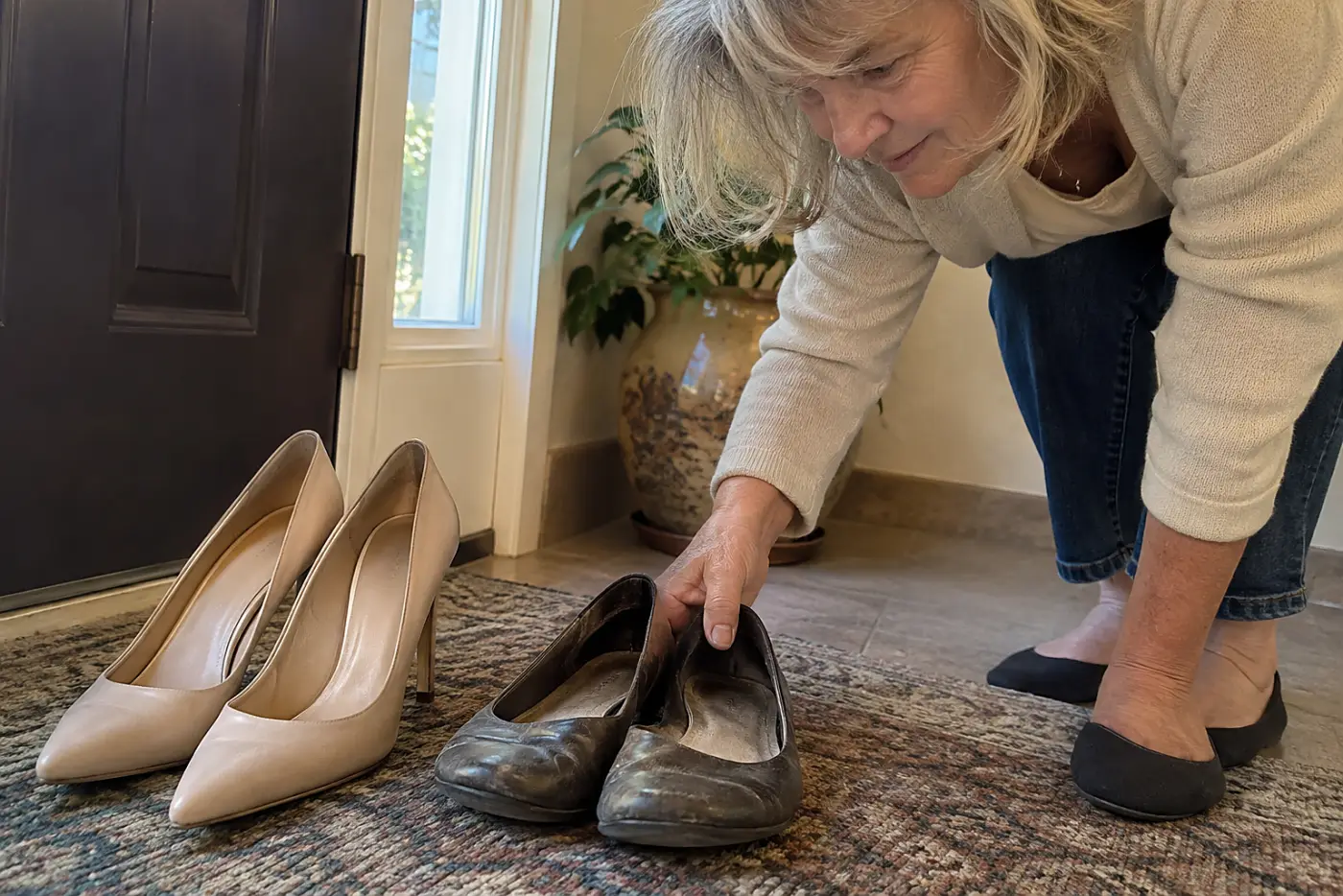Slim heels and broken-in flats sit side by side near the front door as a woman in her sixties reaches for the flats.