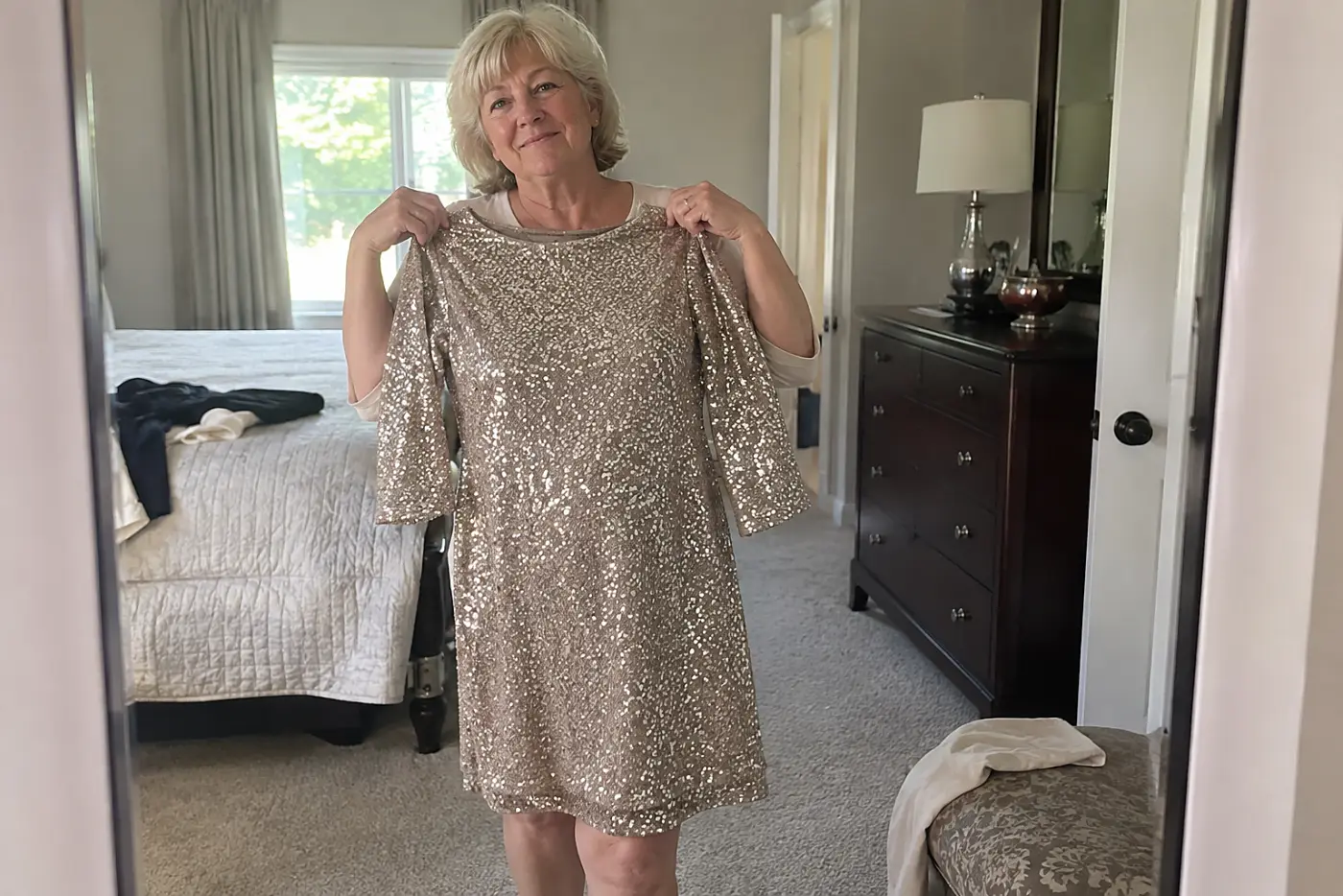 A woman in her sixties holds a sequin dress against herself in a bedroom mirror.