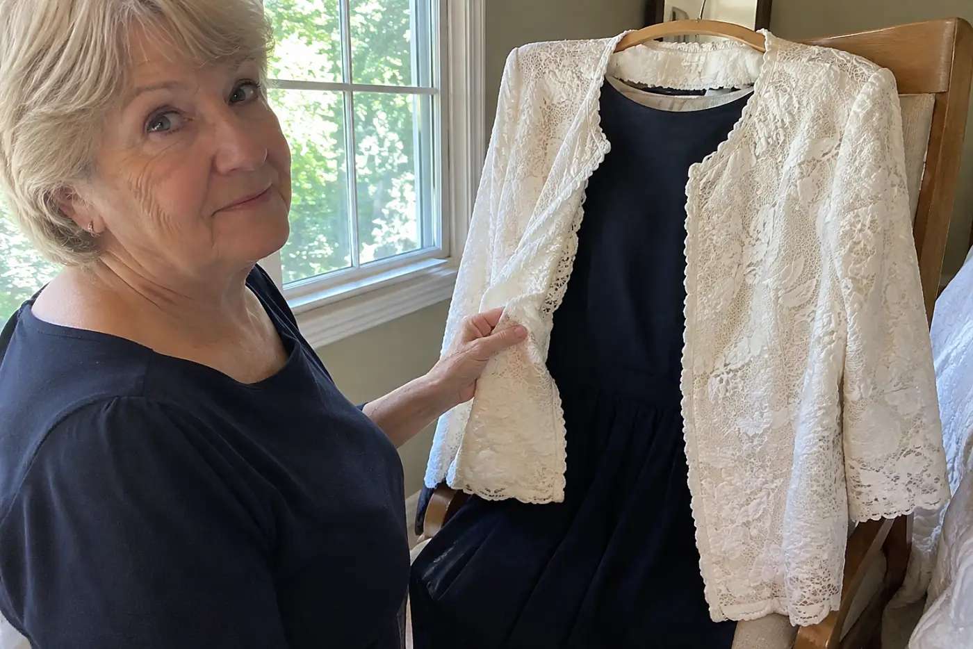 A white lace jacket hangs over a chair beside a dark dress in a real bedroom as a woman in her sixties considers both.