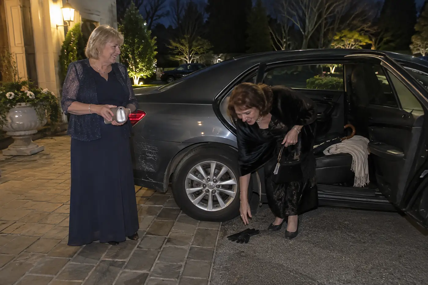 A woman in a navy dress watches another reaching for dropped gloves beside a car.